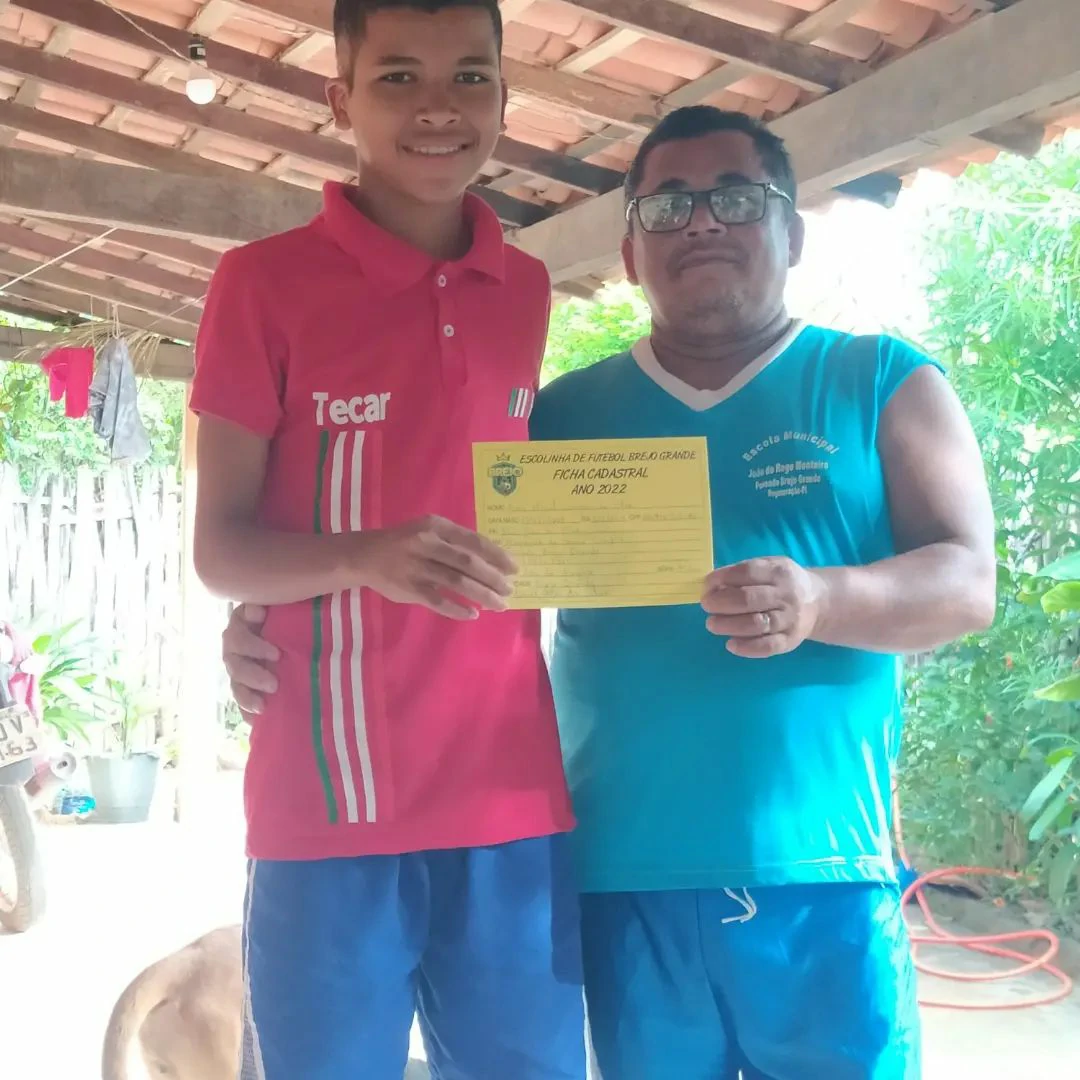 Na - imagem - uma - criança- ao - lado - do – pai - exibindo - com - orgulho - a - ficha - de inscrição - da - Escolhia - de - Futebol – do - Brejo - Grande 