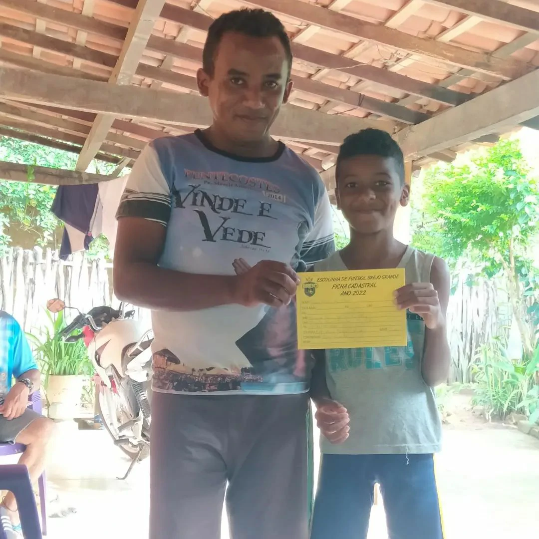 Na - imagem - uma - criança- ao - lado - do – pai - exibindo - com - orgulho - a - ficha - de inscrição - da - Escolhia - de - Futebol – do - Brejo - Grande 